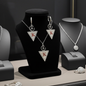 A jewelry display features the Collier LALLA silver necklace and matching earrings, both with large triangle pendants showing a red K and circle-cross symbols, elegantly arranged on a black stand with other elegant pieces.